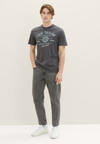 Grå t-shirt med "TOM TAILOR" tekst, korte ærmer, rund halsudskæring, parret med grå jeans og hvide sneakers på en neutral baggrund.