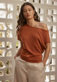 Un pull rouille, décolleté asymétrique, fait d'un tissu léger et texturé, associé à un pantalon beige à coupe décontractée.