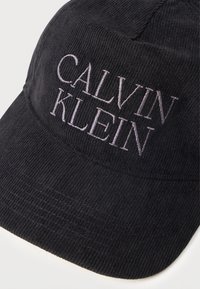 Czarna czapka z kordurowego materiału z haftowanym logo "CALVIN KLEIN" w kolorze szarym. Strukturalny kształt z zakrzywionym daszkiem i teksturowanym materiałem.