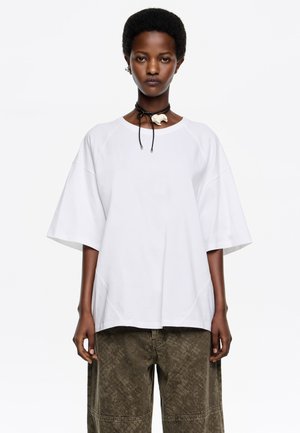 Biała, oversize'owa koszulka z szerokimi rękawami i okrągłym dekoltem, w zestawieniu z oliwkowozielonymi, teksturowanymi spodniami. Modelka nosi czarną chokerową naszyjnik.