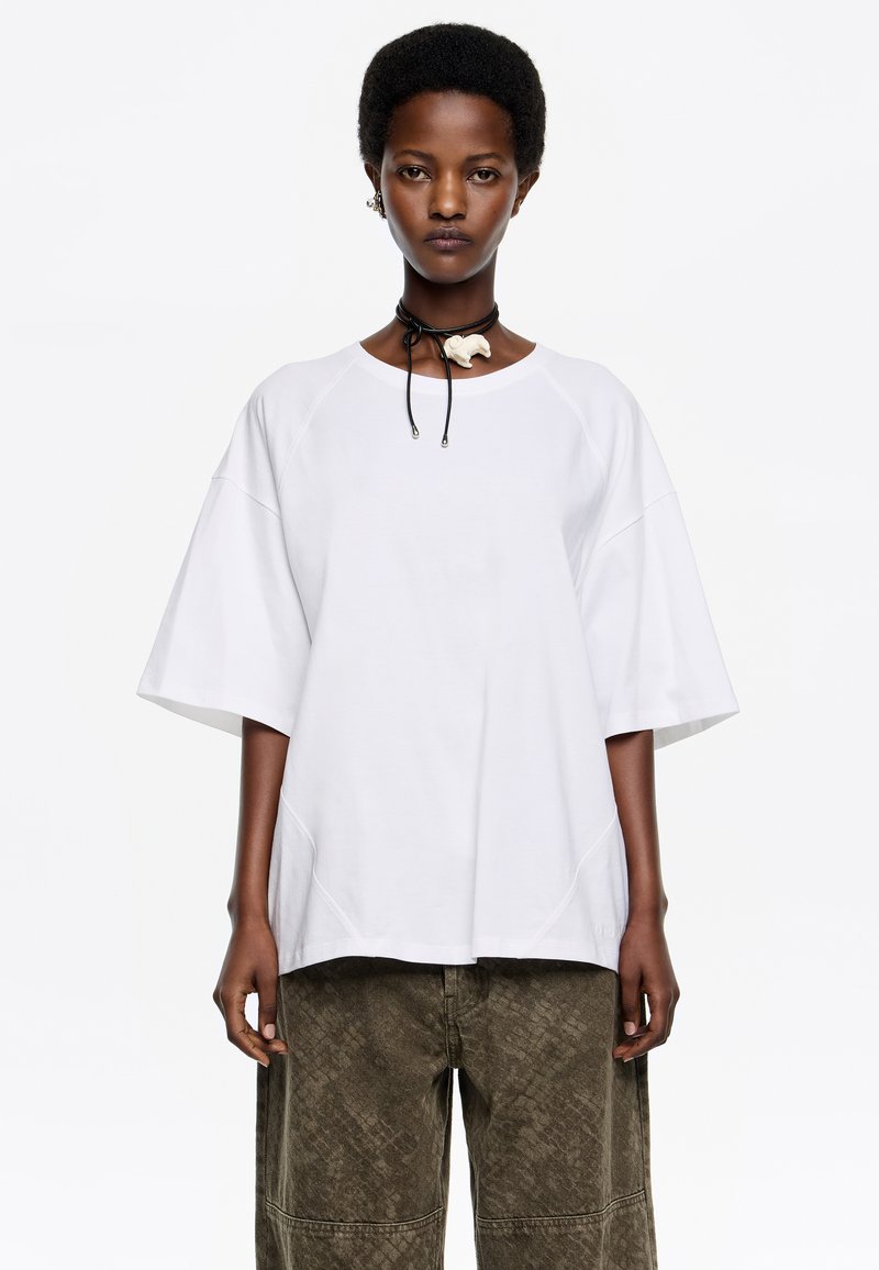 Camiseta blanca de gran tamaño con mangas anchas y cuello redondo, combinada con pantalones de textura verde oliva. La modelo lleva un collar choker negro.