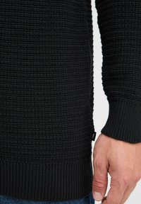 Schwarzer Strickpullover mit strukturiertem Muster, gerippten Bündchen und einem Seitenschild. Der Stoff hat ein dichtes, warmes Erscheinungsbild.