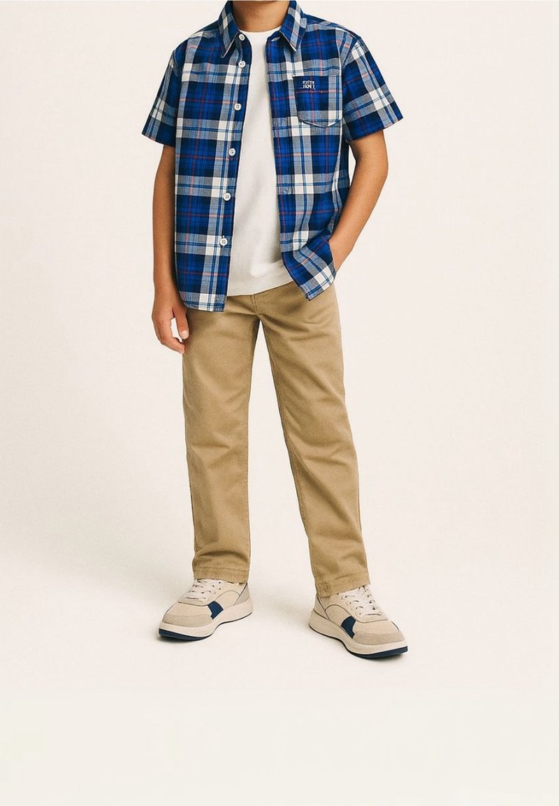 Garçon portant une chemise à manches courtes à carreaux bleus par-dessus un t-shirt blanc, un pantalon beige et des baskets blanches avec des accents bleu marine, debout avec une main dans la poche.