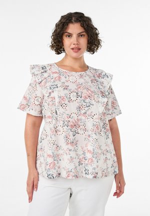 Kvinde med krøllet hår iført en hvid blomstret bluse med flæse skuldre og hvide bukser, stående mod en ensfarvet baggrund.
