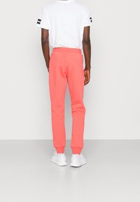 Person som bär korallrosa joggers, vita sneakers och en vit t-shirt med korta ärmar och svarta ränder på ärmarna, stående med ryggen mot kameran.