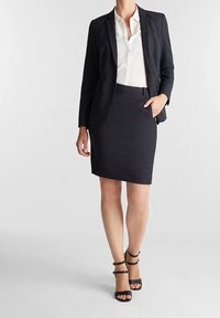 Blazer bleu marine avec une texture subtile, associé à une jupe ajustée. Blouse blanche en dessous. Sandales à talons hauts noires à lanières complètent le look.