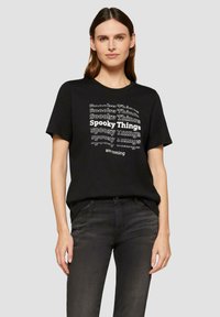 T-shirt noir avec un design en texte blanc répété : "Choses Effrayantes", avec des styles de police variés. Manches courtes, col rond, coupe décontractée.