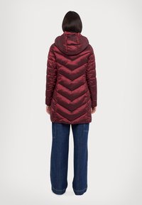Burgundy puff jacket dengan penutup kepala, menampilkan pola chevron, tekstur mengilap, dan desain panjang menengah. Dipadukan dengan celana denim berpotongan lebar.