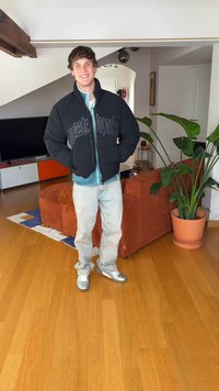 Juoda pūkinė striukė su logotipu, po ja šviesiai mėlyni marškiniai, šviesūs džinsai ir sidabriniai sportbačiai. Vidaus aplinka su augalu ir sofa.