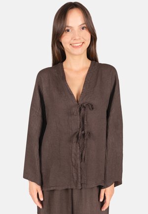 Junge Frau trägt ein lockeres braunes Langarmshirt mit zwei vorne gebundenen Knoten, lächelnd vor einem schlichten hellen Hintergrund.