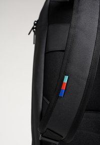 Sac à dos noir en tissu durable avec une surface texturée. Dispose d'une sangle réglable avec une petite étiquette colorée (rouge, bleu et aqua).
