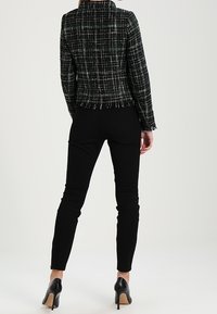 Donna che indossa una giacca fitted a quadri nera, pantaloni slim neri e scarpe con tacco alto nere, in piedi con le spalle alla fotocamera su uno sfondo bianco.