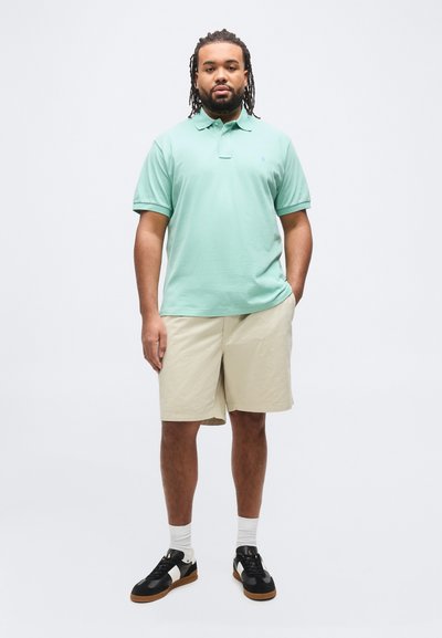 Άνδρας με dreadlocks που φοράει πολο πουκάμισο σε μέντα πράσινο, μπεζ σορτς, λευκές κάλτσες και ασπρόμαυρα αθλητικά παπούτσια, στέκεται με το ένα χέρι στην τσέπη.