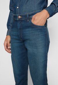 Jean en denim bleu foncé avec une coupe slim. Les caractéristiques incluent une fermeture à bouton à l'avant, cinq poches et des coutures orange contrastantes.