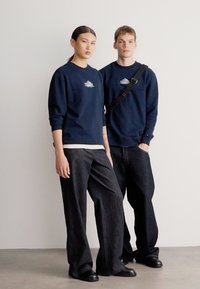 Tengerészeti pulóverek logóval, laza sötét farmernadrággal párosítva. Mindkettőn fekete cipő. Egyszerű, hétköznapi városi öltözet. Minimalista háttér.