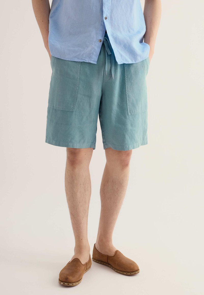 Personne portant un short bleu clair à cordon, une chemise bleu clair boutonnée et des chaussures marron sans lacets sur un fond uni.
