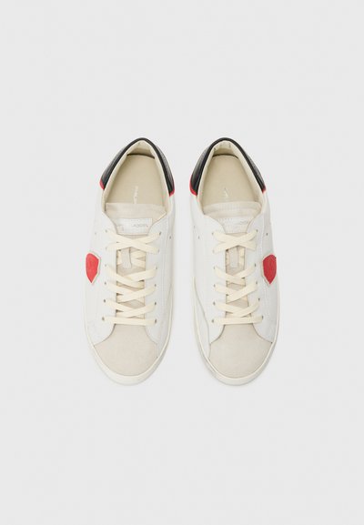 Baskets en cuir blanc avec un accent en forme de cœur rouge et des bordures de talon noires, dotées de bouts en daim beige et de lacets plats beiges.