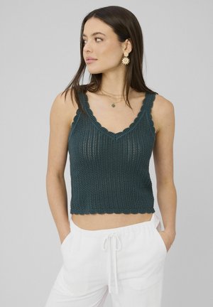 Femme portant un haut sans manches en crochet bleu sarcelle foncé et un pantalon blanc à cordon, regardant sur le côté sur un fond gris uni.