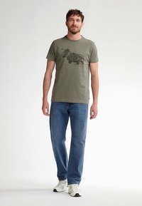 Oliivinvihreä lyhythihainen puuvillainen t-paita, jossa on kuorma-auton graafinen printti. Yhdistetty sinisiin suoralahkeisiin farkkuihin ja valkoisiin lenkkareihin.