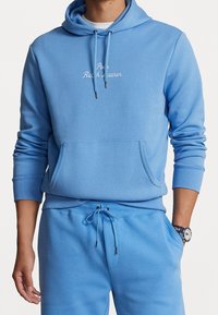 Person som bär en ljusblå huvtröja och matchande sweatpants med "Polo Ralph Lauren" broderat på bröstet och en armbandsur på vänster handled.