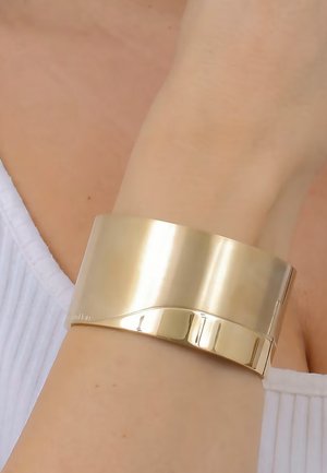 Armbanduhr aus goldfarbigem Metall mit einer glatten, glänzenden Oberfläche und einem subtilen Wellenmuster. Verfügt über einen Scharnierverschluss für einen sicheren Sitz.