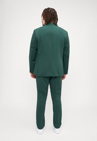 Zelený strihový oblek pozostávajúci z saka a nohavíc, vyrobený z hladkej látky. Obsahuje minimalistický dizajn a je nosený s bielymi teniskami.
