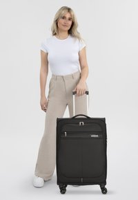 Valigia nera con ruote, dal rivestimento testurizzato, tasca frontale con zip e manico. Il modello è in piedi accanto ad essa, indossando una maglietta bianca e pantaloni beige.