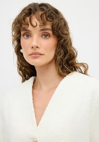 Chaqueta blanca texturizada con un escote en V profundo, que presenta un solo botón decorativo y una tela suave. La modelo tiene el cabello ondulado y un maquillaje ligero.