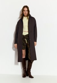 Lang brun frakke med stor krave, lukket foran, lagd over en lys grøn ribbet crop top, parret med ternede shorts og brune knæhøje støvler.