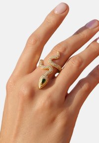Bague en or en forme de serpent ornée d'une pierre précieuse verte et de petites pierres transparentes qui accentuent le design. Portée sur une main avec un vernis à ongles neutre.