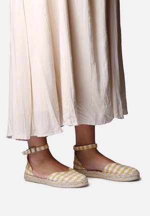 Feet wearing yellow and white gingham espadrilles with ankle straps, paired with a flowing off-white midi skirt.