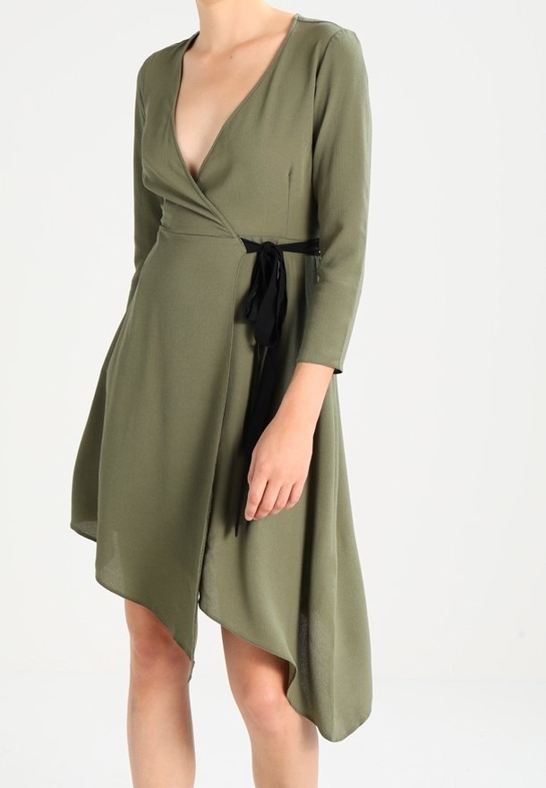 Femme portant une robe portefeuille vert olive à manches trois-quarts et ourlet asymétrique, nouée à la taille avec un ruban noir.