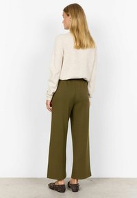 Woman with straight blonde hair wearing a beige sweater, olive wide-leg pants, and dark brown studded loafers, standing against a white wall.