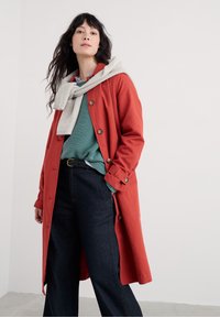 Roter Trenchcoat mit Knöpfen, drüber ein strukturiertes tealfarbenes Pullover und kombiniert mit dunklen Jeanshosen, dazu ein hellgrauer Schal.