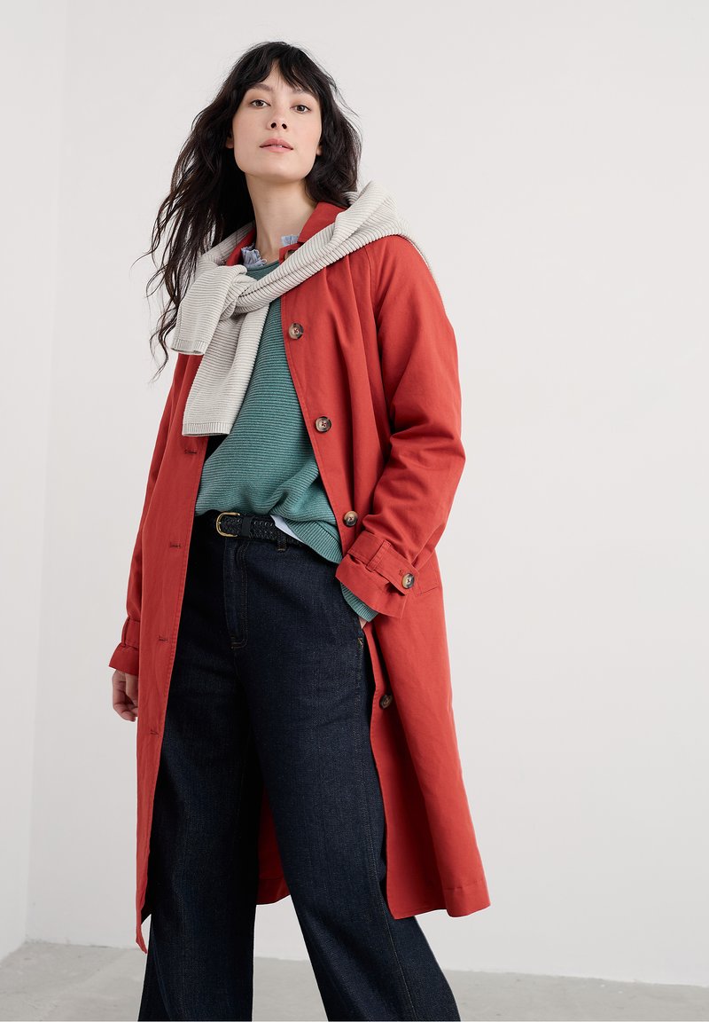 Roter Trenchcoat mit Knöpfen, drüber ein strukturiertes tealfarbenes Pullover und kombiniert mit dunklen Jeanshosen, dazu ein hellgrauer Schal.