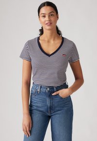 Camiseta de cuello en V de manga corta a rayas en azul marino y blanco con un pequeño logo rojo, combinada con jeans azules de cintura alta, en un fondo simple.
