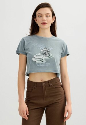 Joven mujer con un top corto gris con estampado de cupcake y texto, combinado con pantalones marrones de talle alto, de pie frente a un fondo liso.