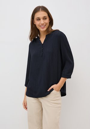 Navyblaues Langarmshirt mit strukturiertem Finish, V-Ausschnitt, drei Knöpfen und lässigem Schnitt, kombiniert mit hellbeigen Hosen.