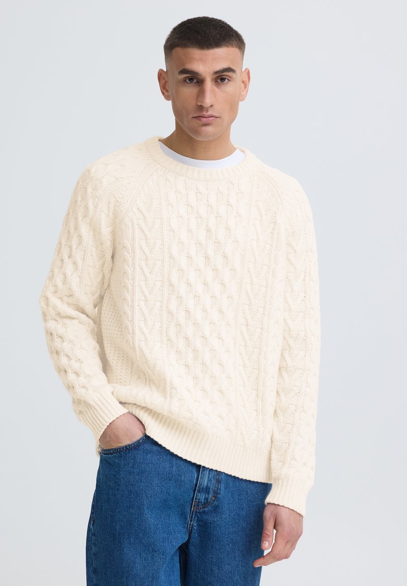 Jeune homme aux cheveux courts portant un pull en maille torsadée crème et un jean bleu, debout, une main dans la poche, devant un fond uni.