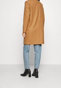 Lange camelcoat met strakke lijnen, gemaakt van een wolmix, gedragen over lichtblauwe jeans, gecombineerd met zwarte enkellaarsjes met hak.