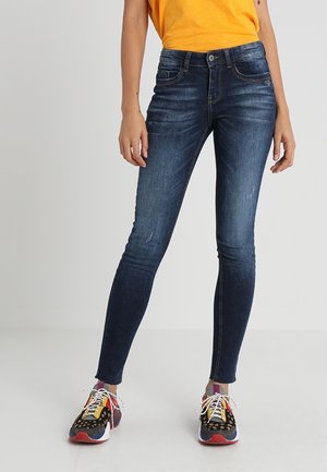 Schmal geschnittene dunkelblaue Jeans, kombiniert mit bunten Sneakers und einem leuchtend orangefarbenen Oberteil, dargestellt vom mittleren Oberkörper bis zu den Füßen vor einem einfarbigen Hintergrund.