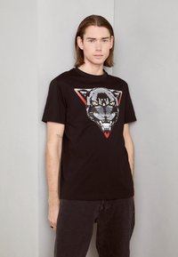 Zwart katoenen T-shirt met een afbeelding van een grommende panter binnen een rode driehoekige omtrek, korte mouwen en een ronde hals.