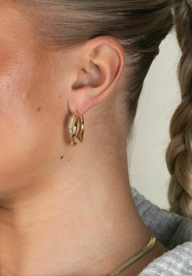 Guldörhängen med en texturerad yta och sammanflätad design. Örhängena är fästa vid loben, vilket framhäver deras cirkulära form.