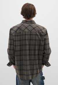 Camicia in flanella a quadri verdi e marroni con maniche lunghe, colletto a punta e orlo curvo. Presenta motivi a quadri contrastanti in tutto il tessuto.