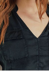 Camicia in tessuto nero e setoso con scollatura a V, dotata di taschino sul petto e dettagli cuciti. Texture liscia e leggera lucentezza.