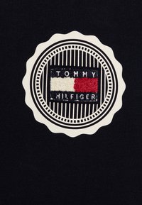 Il tessuto nero presenta un logo circolare di Tommy Hilfiger con un rettangolo ricamato in rosso, bianco e blu, delimitato da un bordo ondulato color crema.