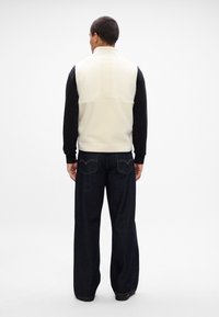 Γουστόζικο γιλέκο με χρωματικό μπλοκάρισμα που έχει κρέμ μπροστινό μέρος και μαύρα μανίκια, φτιαγμένο από μαλακό ύφασμα. Συνδυασμένο με σκούρα μπλε παντελόνια σε χαλαρή γραμμή και μαύρα παπούτσια.