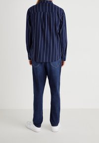 Camisa de botón a rayas azul marino con diseño en la parte trasera, combinada con jeans de un azul oscuro y zapatillas blancas. Textura de tela suave.