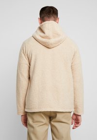 Beige fleece hoodie med en blød, tekstureret yderside, forsynet med hætte og lige kant, båret over lysfarvede bukser.