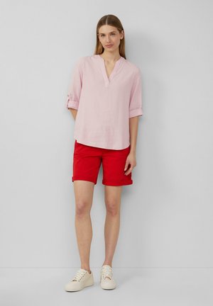 Blouse en lin rose clair avec un col en V et des manches retroussées, associée à un short rouge roulé et des baskets beiges. Tenue simple et décontractée.